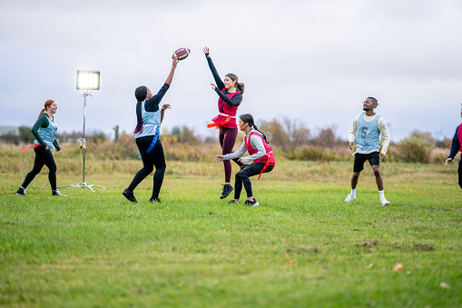 A group of University students run around on a field as they play a game of flag football together.  They are each dressed comfortably and have numbered team pinnies on.