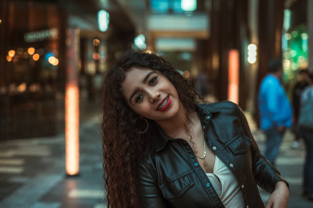 Brunette girl with curly hair enjoys a moment of lights while artificial as night falls, she wears a black leather jacket.