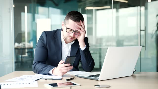 Disappointed stressed businessman in formal suit reads bad news on smartphone sitting at workplace
