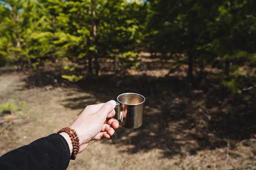 A man's hand holds a mug of tea against the background of the forest, a metal mug for coffee to drink on a hike, dishes in the forest, nature pine sun, rudraksha bracelet. High quality photo