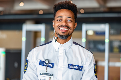 Portrait of a mid adult Black male security worker posing for a photo in an airport lobby, looking at the camera, wearing a uniform shirt and an ear piece.