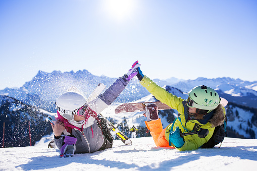 two female friends with ski and snowboard equipment having fun and high five with snow in the air.