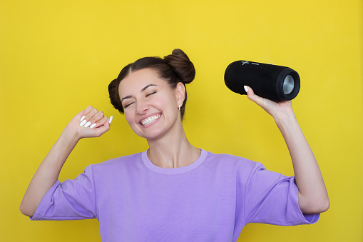 a young pretty Caucasian woman in a purple T-shirt listens to music and dances, closing her eyes, holding a wireless music speaker in her hand. Isolated on a yellow background. Copy space