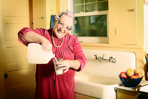 A granny in curlers and cat's eye glasses in her kitchen near the sink pouring a glass of milk. More granny images.