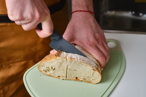 Man cuts a piece of fresh bread with a knife and makes a delicious cheese sandwich in the kitchen