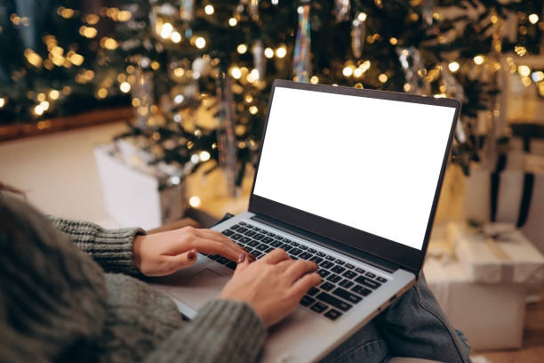 istock woman work on laptop computer with blank display screen with mockup copy space. home interior decorated for Christmas celebration with Christmas tree and garland lights. 1767592574