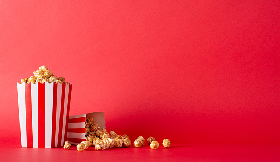 Host movie premieres with your buddies and delicious snacks. Side view photograph of tabletop featuring mouthwatering popcorn in striped boxes against red wall, providing space for movie advertisement