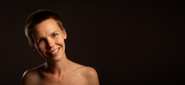 A woman in her prime, donning a short hairstyle and baring her shoulders, wears a gentle smile, positioned against a black background with an empty banner for your text needs