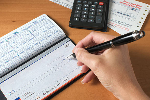 Writing a check to pay the bills, hand holding pen "Woman's hand writing a check to pay the bills, with calculator and an invoice on the desktop. Generic account numbers and bank info." financial item stock pictures, royalty-free photos & images