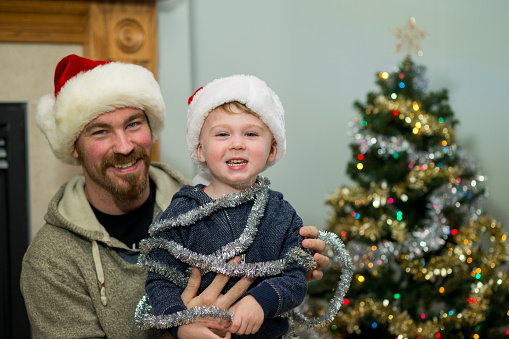 Cute little boy wearing a festive Santa hat is laughing as his father has wrapped him in tinsel as they are decorating the Christmas tree together as part of a Christmas Eve tradition as a family.