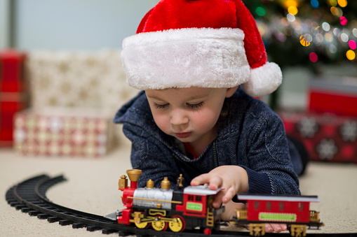 Cute little boy in a festive red and white Santa Clause hat is laying on his stomach as he is playing with a toy train he received as a gift on Christmas morning in front of the decorated Christmas tree.