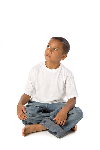 A mixed race little boy sits cross-legged and looks up as if thinking.