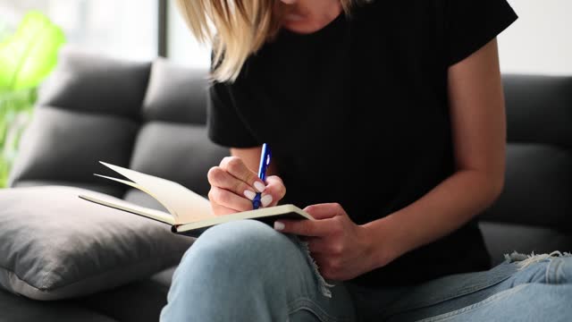 Woman writing notes with ballpoint pen in notepad on sofa at home closeup 4k movie