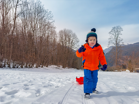 Little boy in winter outfit pull red sledge mounting the slope at mountain vacation
