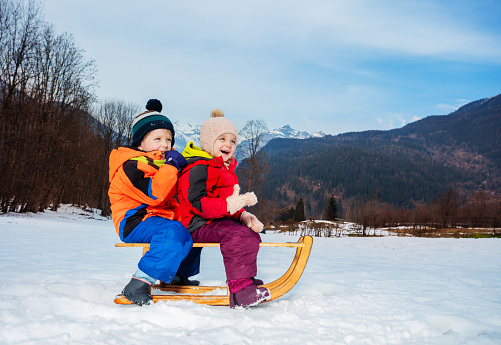 Laughing little boy with girl in winter coats and woolen hats siting on the sledge on the snow smiling