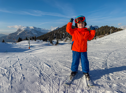 Little kid skier, pupil of ski school, standing on slope, making victory gestures in helmet and mask ready to ride in French sunny mountains