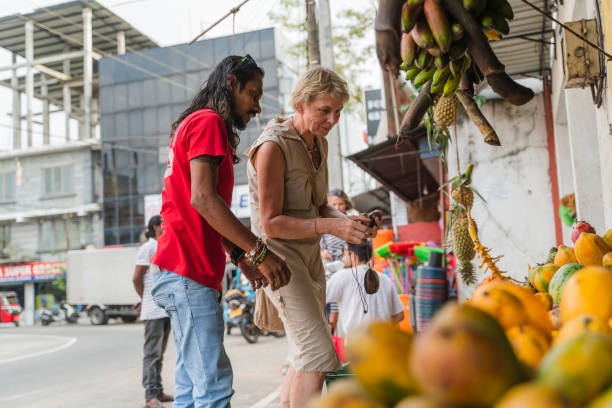 ein mann aus sri lanka hilft einer europäischen touristin, einer 50-jährigen aktiven frau, auf einem lokalen straßenmarkt in aluthgama, sri lanka, frische, reife früchte zu pflücken. - 30s women asian ethnicity 35 40 years stock-fotos und bilder