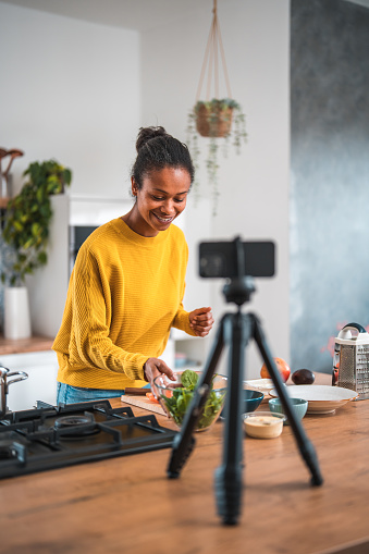 A dark-skinned woman creating deliciously healthy dishes in her captivating food vlog. Using a mobile phone camera, she filming herself in her domestic kitchen.