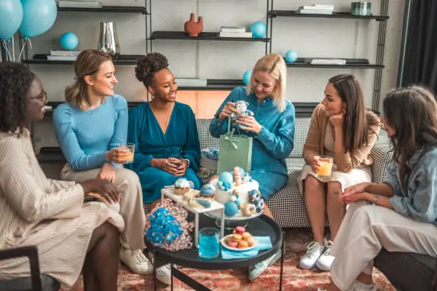In a cozy living room, a group of diverse best friends gifts their pregnant blonde friend while enjoying each other's company. The room is decorated with baby shower decor, cakes, and sweets.
