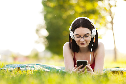 Beautiful dark hair girl listening to music with a headphones from smartphone. Lying on a green meadow.