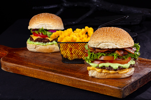 2 giant hamburgers with an iron basket containing fries, on top of a wooden board and all on a table with black fabric and a black background