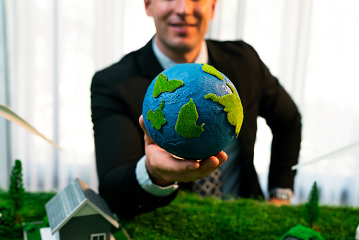 Eco business and save Earth concept shown by businessman or CEO holding paper globe in office with eco friendly mockup to promote CO2 and carbon footprint reduction for greener future. Quaint