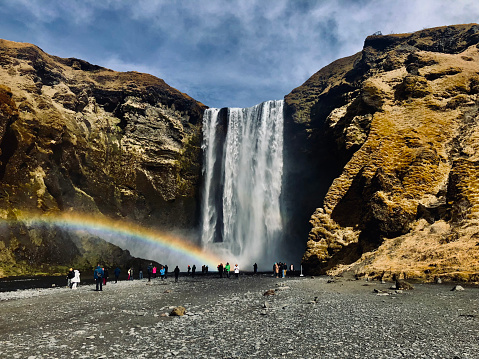Skogfoss waterfall in Iceland. The water is falling from a cliff down to a river. There is grass growing and gravel on the path. There is a rainbow and the sky is blue with clouds.