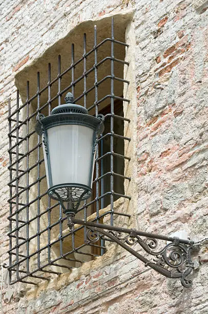 Medieval window with grating on an ancient brick wall. In foreground an old-looking public streetlamp.