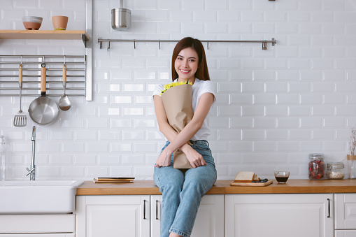 Portrait of beautiful feminine Asian woman holding yellow flowers and looking at camera while sitting on top counter on modern kitchen. Romantic portrait. Woman gets gift from boyfriend. Lifestyle