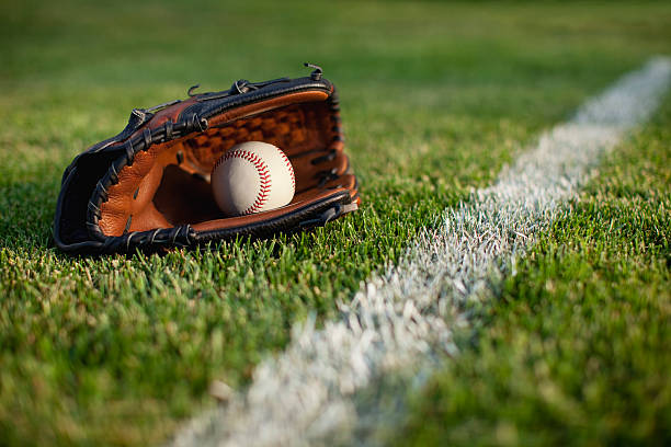 Fictitious baseball stadium full of fans in the stands with baseball glove and ball on infield dirt clay. Deliberate focus on equipment and foreground with shallow depth of field on background. Floodlights flare for effect and copy space. Stadium created in Photoshop.
