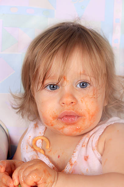 Young blue eyed baby girl making a mess on her face with spaghetti and tomato sauce with pieces in her shirt