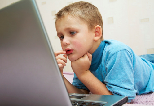 School-age boy sitting in front of the monitor laptop at home on the couch.