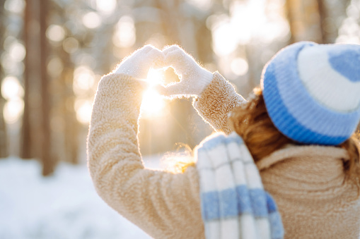 A woman in a hat and scarf against the backdrop of a snowy winter forest holds her hands in the shape of a heart. Back view. Young woman enjoying a sunny winter day. Vacation concept, lifestyle.