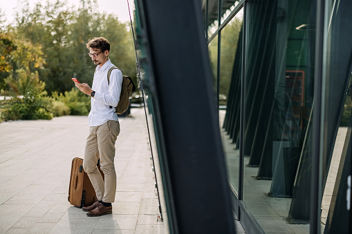 Handsome businessman standing with suitcase outside  and using mobile phone