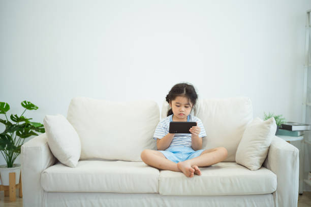 Asian child girl looking using and touch mobile phone screen on couch sofa. Baby smiling funny time to use mobile phone. Too much screen time. Cute girl watching videos while tv, Internet addiction. Asian child girl looking using and touch mobile phone screen on couch sofa. Baby smiling funny time to use mobile phone. Too much screen time. Cute girl watching videos while tv, Internet addiction. asian kid too much screentime stock pictures, royalty-free photos & images