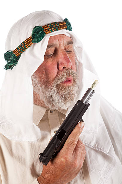 Arab Man Blowing on Money Stuffed in a Gun Older Arabic man in traditional headdress blowing on a hundred dollar bill stuffed in a gun barrel. north african ethnicity stock pictures, royalty-free photos & images