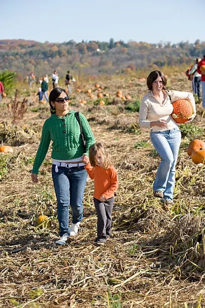 "Lifestyle image, and one of a series: This is a family out on the farm on a pumpkin patch. The mother carries a pumpkin which they will take home and carve. The small girl holds her older sister's hand. There are anonymous people defocused in the background, adding to the bustle of the scene."