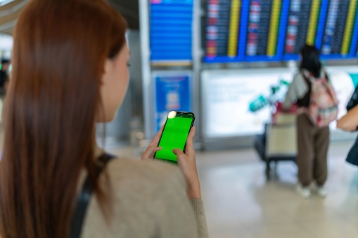 Rear View of Young asian woman carrying handbag and holding smartphone on hand, standing in front the FIDS departure board in airport terminal. Business person on business trip. Young Asian Female Traveler with Smartphone in Airport Terminal.