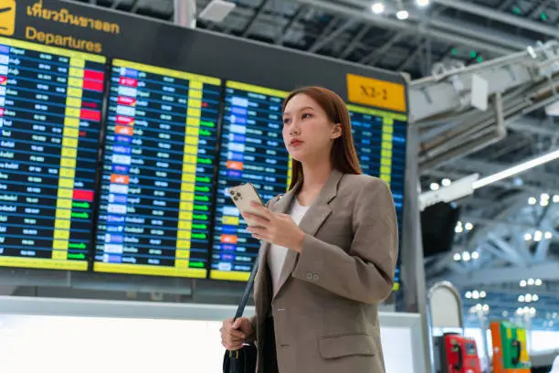 Photo of Young asian woman carrying handbag and holding smartphone on hand, standing in front the flight information display departure board in airport terminal. Business person on business trip. Young Asian Female Traveler with Smartphone in Airport Terminal.