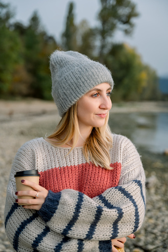 Side view of positive young female long blond hair in warm clothes smiling, looking away while standing on shore of lake, holding coffee, in background trees, lake and mountains. lifestyle concept