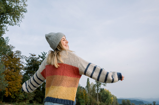 From below of young female in warm sweater and knitted hat standing against green trees and cloud while enjoying sunny day in countryside on winter time