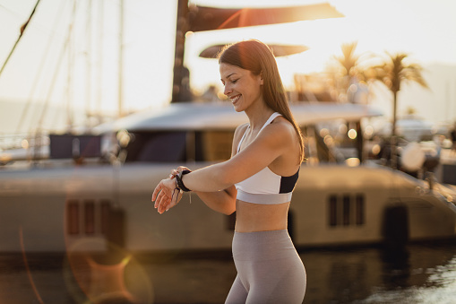 Beautiful young woman wearing sports clothing, exercising, and checking the information on her smartwatch in a seaport on a sunny summer day