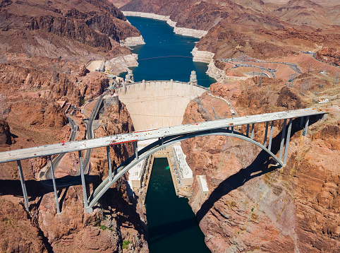 An aerial view of the Colorado River Bridge and the Hoover Dam in Nevada, Arizona, USA.  There is a hydroelectric power station, a bridge and a dam wall.  On either side of the bridge are rock formations that feature roadways.