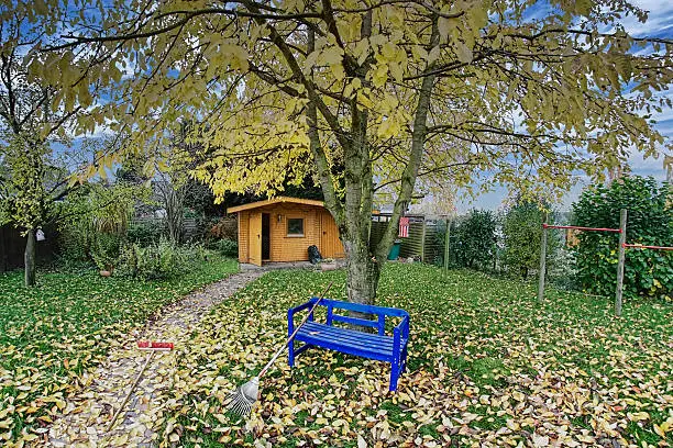 "HDR Image taken with a super wide-angle lens.Garden during fall with a blue bench, red broom to clean the garden-path and a rake for the foliage."