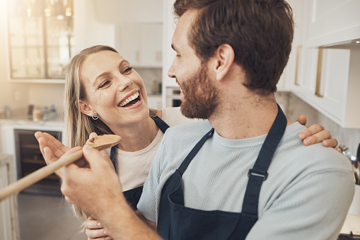 Cooking, taste and happy couple in kitchen bonding while preparing a meal in their home together. Food, spoon and man feeding woman in a house with romance, smile and having fun on weekend or day off