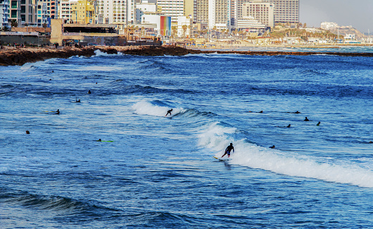 Surfers in distance in Tel Aviv and hotels or residential buildings next  to coastline in Tel Aviv .  2  person rides the wave - blurred motion so unrecognizable persons 
. Hotel or residentoal buildings