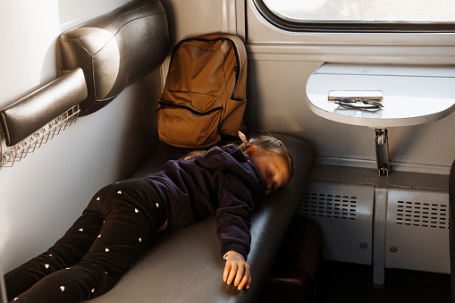 Tired child riding in train on sunset, bright sunlight, atmospheric travel by railway with kids. Passenger resting on the couch of slipping wagon. Long family trip
