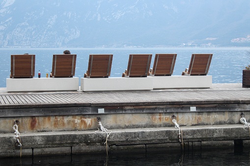 Tourists, day trippers or locals enjoy a spring day on the shores of Lake Garda. They rest on wooden loungers.