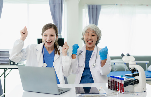 Successful Asian female doctor celebrates victory over successful treatment of patient in hospital, sitting at desk, arms raised with cheerful smile, research concept.