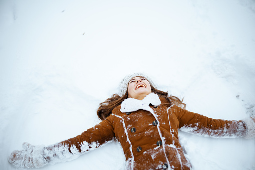 happy modern middle aged woman in brown hat and scarf in sheepskin coat with mittens making snow angels outside in the city in winter.